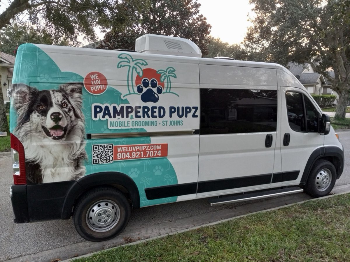 A white Pampered Pupz mobile grooming van parked on a residential street, wrapped in teal with a palm-tree paw logo and a photo of a merle Australian Shepherd